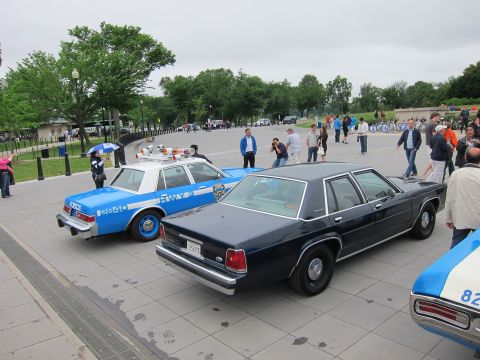1991 Ford LTD Crown Victoria - dark navy Ford sedan parked beside a blue-and-white NYPD police cruiser on a stone plaza with onlookers nearby.