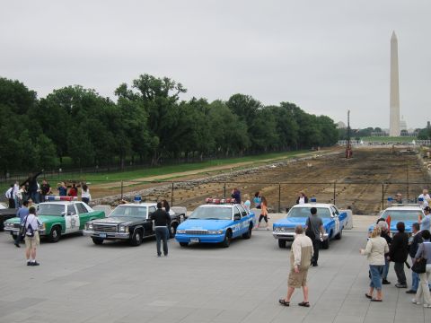 1991 Ford LTD Crown Victoria - Green and blue vintage police cruisers parked on a plaza with the Washington Monument rising in the hazy background.
