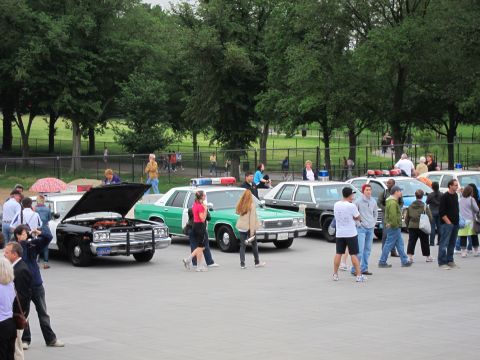 1991 Ford LTD Crown Victoria - black police car with its hood raised sits among green and dark vintage cruisers as visitors mill around the display.