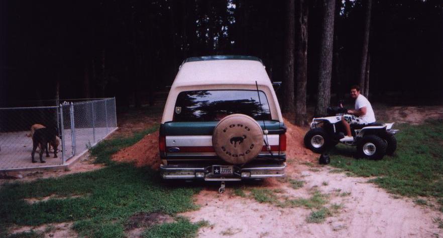 Green Ford Bronco with white top and Ford Bronco spare cover parked on dirt mound in yard