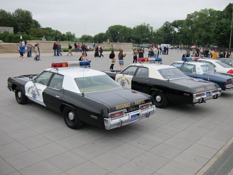 1991 Ford LTD Crown Victoria - Several black-and-white Highway Patrol cruisers parked in a row on a plaza as people walk past in the background.