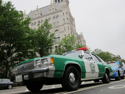 1991 Ford LTD Crown Victoria - Low-angle view of a green-and-white Ford police sedan with star decal parked in front of a tall ornate stone clock-tower building.