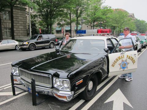 1991 Ford LTD Crown Victoria - woman and a man in a cap stand beside a black-and-white Highway Patrol sedan with light bar, parked on a city street.