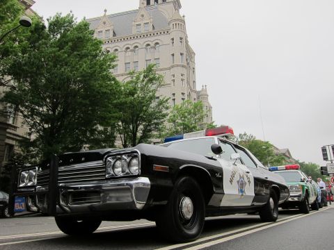 1991 Ford LTD Crown Victoria - Dramatic low-angle front view of a black-and-white police sedan with light bar parked before a tall ornate stone building.