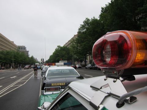 1991 Ford LTD Crown Victoria - Close-up of a red-and-amber emergency rotating light mounted on a police car roof, overlooking a row of parked cars on a city street.