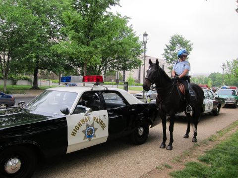 1991 Ford LTD Crown Victoria - mounted police officer on a dark horse stands beside a black-and-white Highway Patrol sedan on a tree-lined sidewalk.