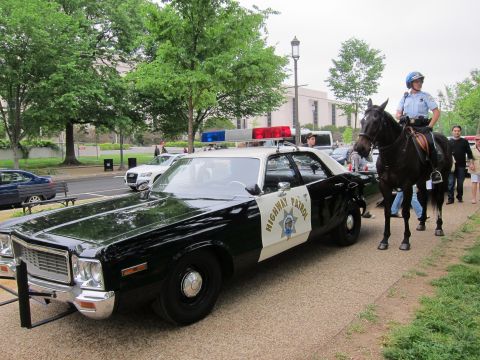 1991 Ford LTD Crown Victoria - black-and-white Highway Patrol sedan with light bar parked at a curb, with a mounted police officer on horseback standing behind it.