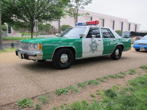 1991 Ford LTD Crown Victoria - green-and-white Ford police sedan with sheriff star decal parked on a gravel path beside a grassy lawn and trees.
