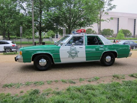 1991 Ford LTD Crown Victoria - Side profile of a green-and-white Ford police sedan marked Sheriff, parked on a gravel path in a tree-lined park.