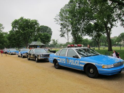 1991 Ford LTD Crown Victoria - blue NYPD-marked Chevrolet Caprice police car heads a row of parked classic patrol cars on a gravel path in a grassy park.