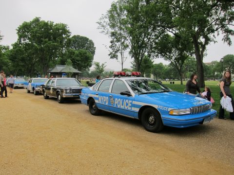 1991 Ford LTD Crown Victoria - blue NYPD-marked police sedan parked at the head of a line of classic patrol cars on a gravel path, with pedestrians nearby.