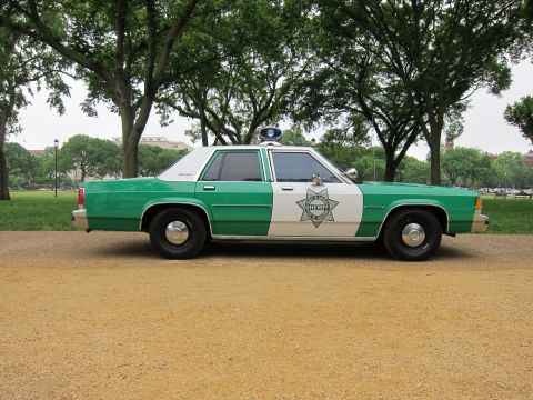 1991 Ford LTD Crown Victoria - Side profile of a green-and-white Ford police sedan with sheriff star decal and roof light, parked on a gravel path in a park.