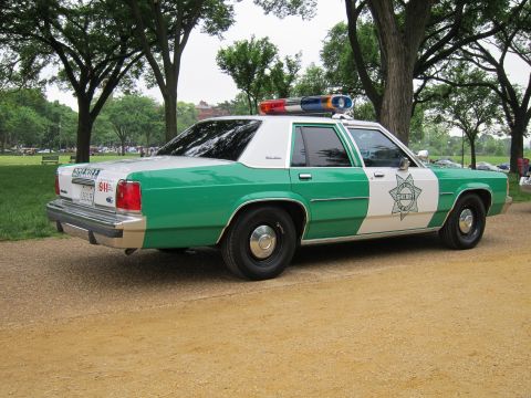 1991 Ford LTD Crown Victoria - Rear three-quarter view of a green-and-white Ford police sedan marked Sheriff with a 911 decal, parked on a park path.