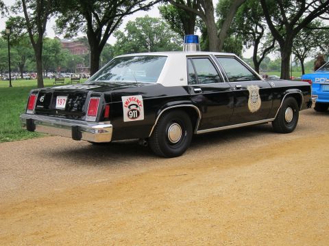 1991 Ford LTD Crown Victoria - black-and-white vintage police sedan with a single roof beacon and a '911 emergency' decal sits on a gravel path in a tree-lined park.