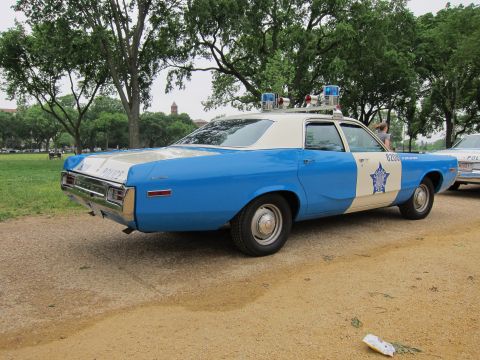 1991 Ford LTD Crown Victoria - blue-and-white vintage police sedan with twin roof beacons and a star badge, seen from the rear with other cars lined up behind.