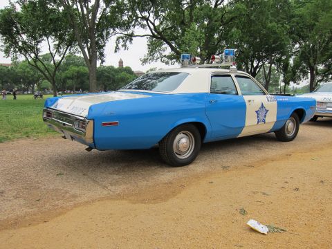 1991 Ford LTD Crown Victoria - blue-and-white vintage police sedan with roof beacons and a star badge, parked on a gravel path in a park.