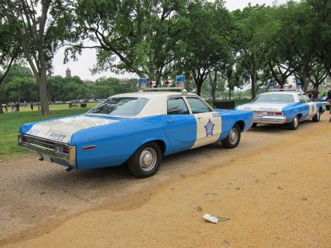1991 Ford LTD Crown Victoria - Two blue-and-white vintage police sedans with roof beacons parked nose to tail along a tree-lined gravel park path.