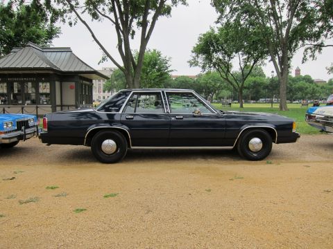 1991 Ford LTD Crown Victoria - dark navy full-size sedan with chrome trim and hubcaps, shown in full side profile on gravel near a park pavilion.