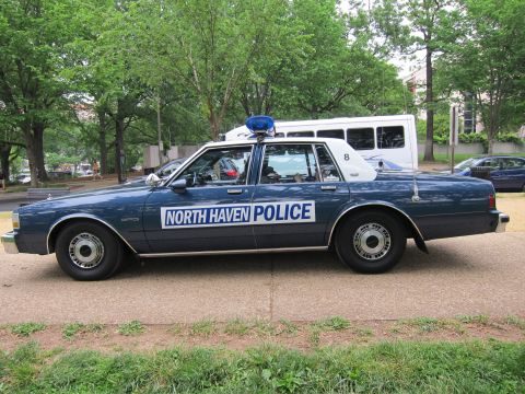 1991 Ford LTD Crown Victoria - dark blue 'North Haven Police' sedan with a single roof beacon, shown in full side profile with a white van in the background.