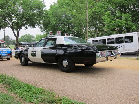 1991 Ford LTD Crown Victoria - black-and-white police sedan with a 'Police' star badge and roof lights, parked on gravel beside an RV in a park.