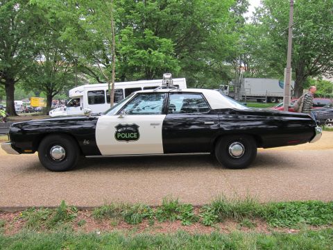 1991 Ford LTD Crown Victoria - black-and-white police sedan with a 'Police' star badge and roof beacon, shown in full side profile on a park path.