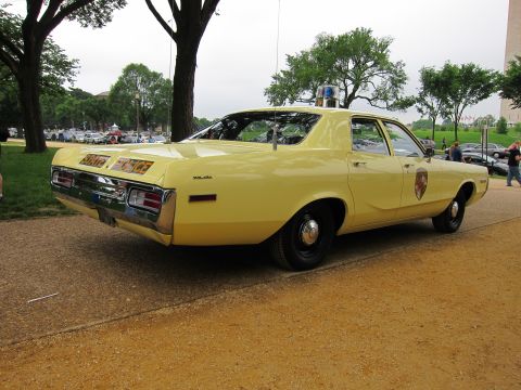 1991 Ford LTD Crown Victoria - pale yellow vintage police sedan with a shield emblem and roof beacon, seen from the rear on gravel in a park.
