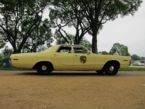 1991 Ford LTD Crown Victoria - pale yellow vintage police sedan with a shield emblem on the door and a roof beacon, shown in full side profile in a park.