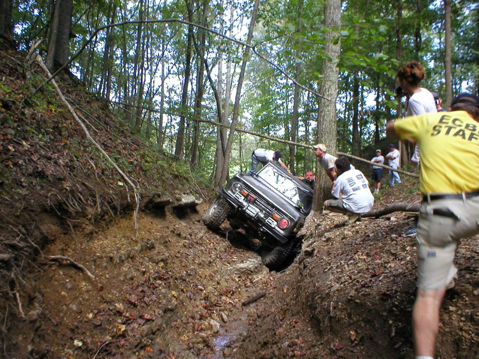 Dark Ford Bronco tackling a steep muddy trail obstacle in the woods as spectators watch