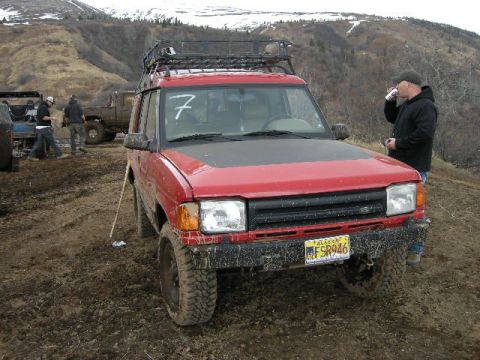 1990 Chevrolet Suburban - This red SUV, likely a 1994 Land Rover Discovery, is equipped for off-road use and is seen in a muddy environment.
