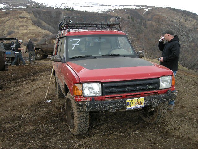 Red off-road SUV with a roof rack and number 7 on the windshield, parked on a muddy hill