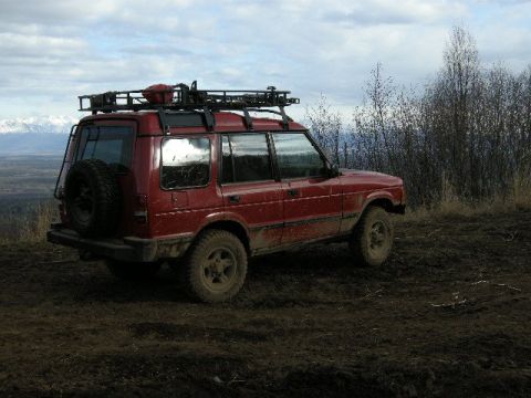 1990 Chevrolet Suburban - A red SUV equipped with a roof rack and external spare tire is seen parked on a muddy, off-road trail.