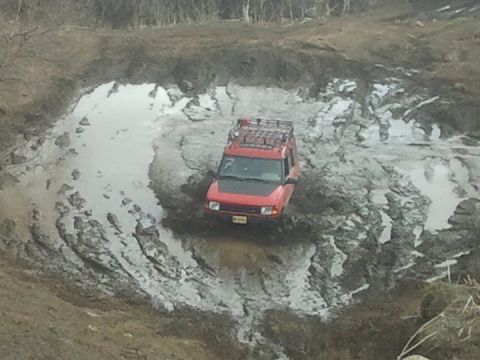 1990 Chevrolet Suburban - This red SUV is navigating a challenging muddy terrain, with its front tires submerged in water.