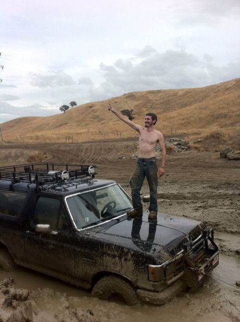 1990 Ford Bronco - A man stands atop a mud-splashed Ford Bronco, stuck in a muddy terrain, pointing towards the sky.