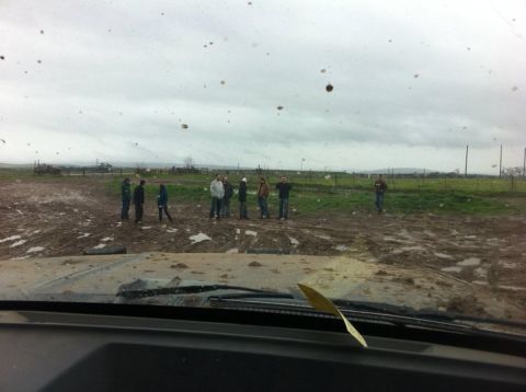 1990 Ford Bronco - A group of people stand in a muddy field under an overcast sky, viewed from a vehicle's interior.
