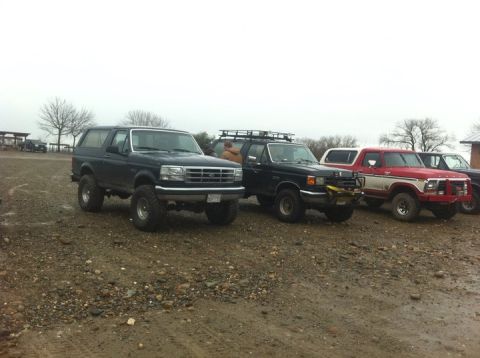 1990 Ford Bronco - Several trucks and SUVs are parked on a muddy lot under an overcast sky.