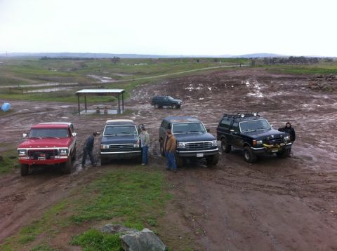 1990 Ford Bronco - Several SUVs are gathered on a muddy terrain under an overcast sky.