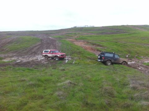 1990 Ford Bronco - A red and white 1990 Ford Bronco assists a black SUV that is stuck in the mud on a grassy hill.