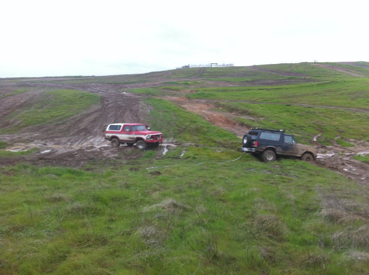 Red and white 1990 Ford Bronco towing a stuck black SUV in muddy terrain