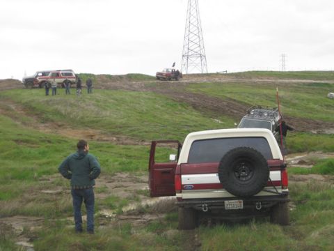 1990 Ford Bronco - Group of Ford Broncos and other SUVs off-roading in a muddy, grassy area with a large power line tower in the background.