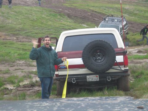 1990 Ford Bronco - A man waves enthusiastically while standing next to a Ford Bronco that appears to be stuck in muddy terrain.