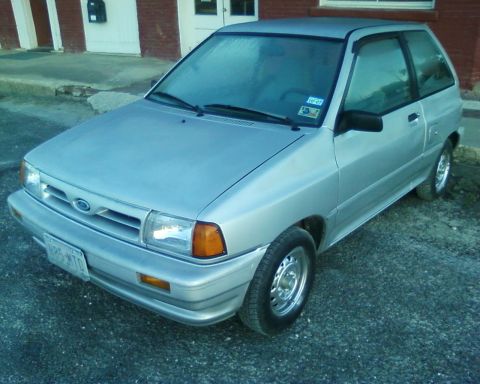 1991 Ford Festiva - A silver 1991 Ford Festiva hatchback is parked outside a brick building. The car is viewed from the front three-quarter angle.