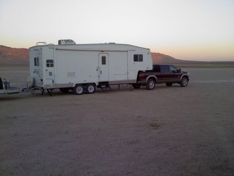 2011 Ford F-250 - A maroon Ford F-250 Super Duty truck is seen from the side, towing a large white fifth-wheel trailer across a barren, dusty terrain.