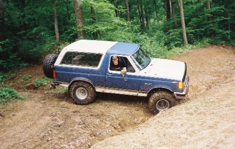 1990 Ford Bronco - A 1990 Ford Bronco navigates a muddy off-road trail surrounded by trees.