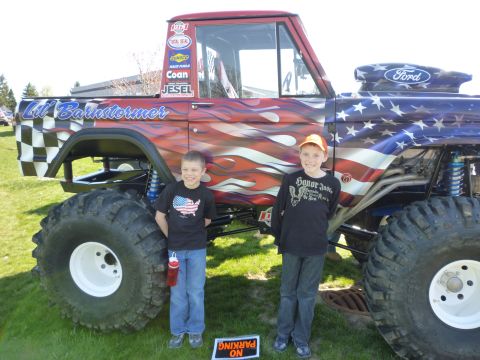 1995 Ford Bronco - A custom monster truck with patriotic paint and flames is shown with two young boys posing next to it.