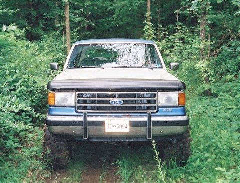 1990 Ford Bronco - A blue and white 1990 Ford Bronco is parked in a wooded area, viewed from the front.
