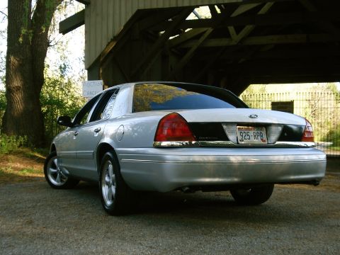 2000 Ford Crown Victoria - Silver Ford Crown Victoria sedan photographed low from the rear three-quarter driver's side on gravel in front of a weathered wooden barn,…
