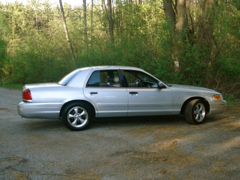 2000 Ford Crown Victoria - A silver Ford Crown Victoria full-size sedan shown in a full side profile view, parked on a gravel lot in front of dense green forest. It h…
