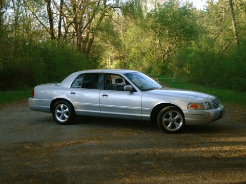 2000 Ford Crown Victoria - Silver Ford Crown Victoria sedan, side profile, parked on gravel beside a wooded area with green spring foliage.