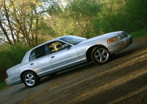 2000 Ford Crown Victoria - A silver Ford Crown Victoria full-size sedan shown from the passenger side at a strong diagonal tilt, parked on a gravel road with green tr…