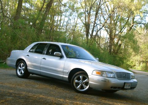 2000 Ford Crown Victoria - A silver Ford Crown Victoria full-size sedan photographed from the front three-quarter angle on a gravel road, backed by dense green trees.…